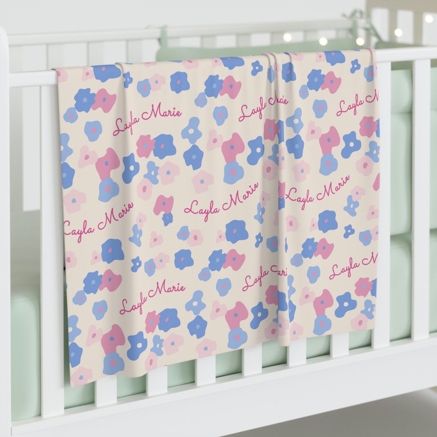 Jersey personalized baby blanket in graphic pink and blue daisy pattern hung over side of white crib