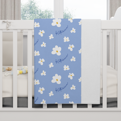 Fleece personalized baby blanket in blue daisy pattern hung over side of white crib with window in the background