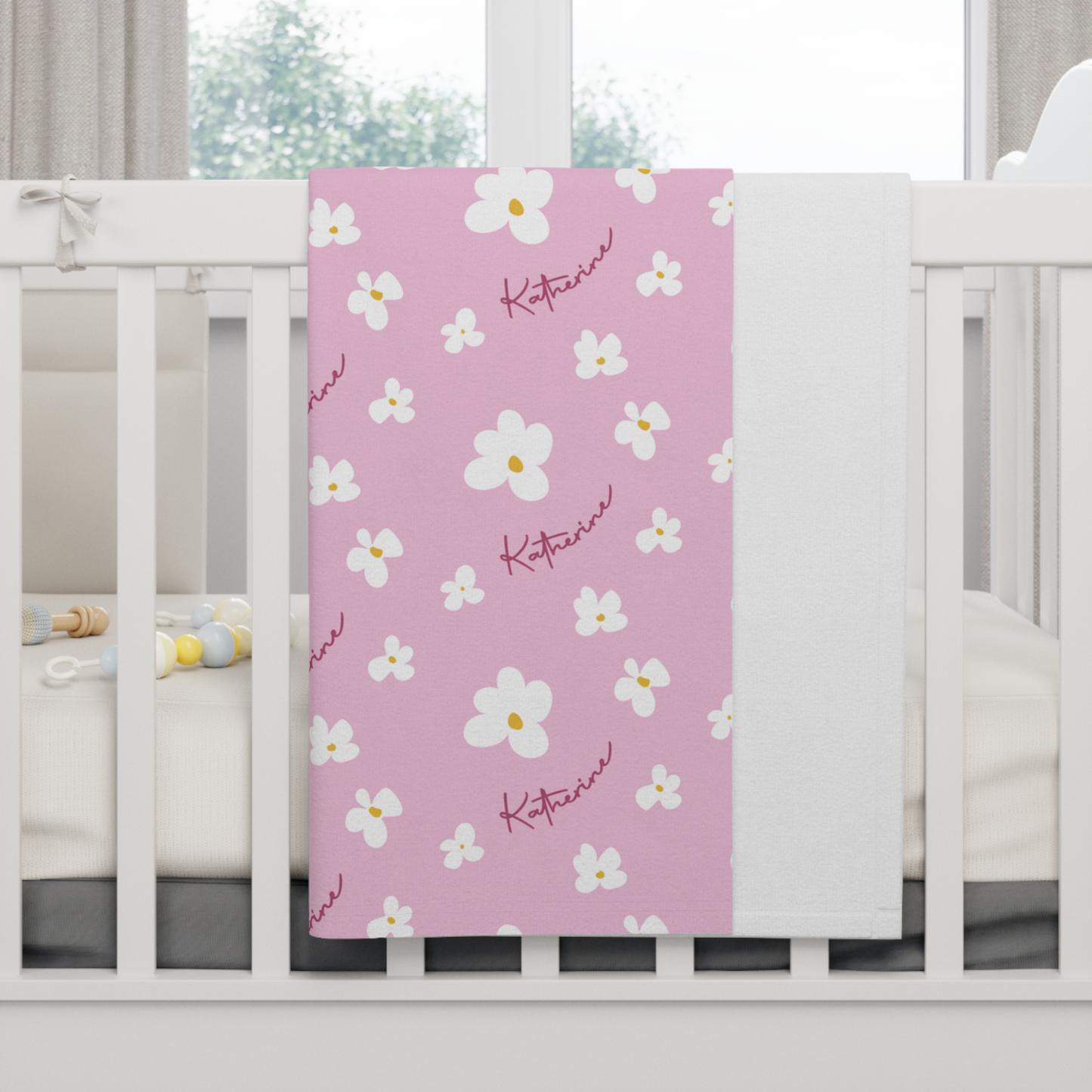 Fleece personalized baby blanket in pink daisy pattern hung over side of white crib with window in the background