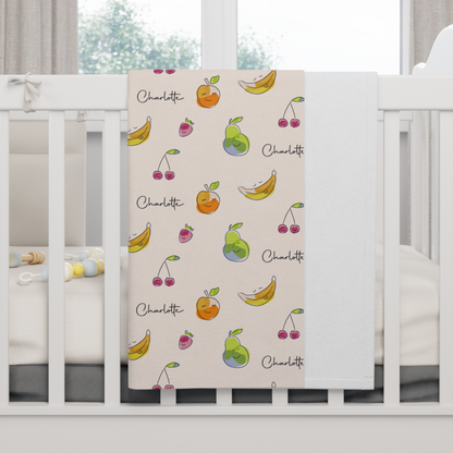 Fleece personalized baby blanket in happy fruit pattern hung over side of white crib with window in the background