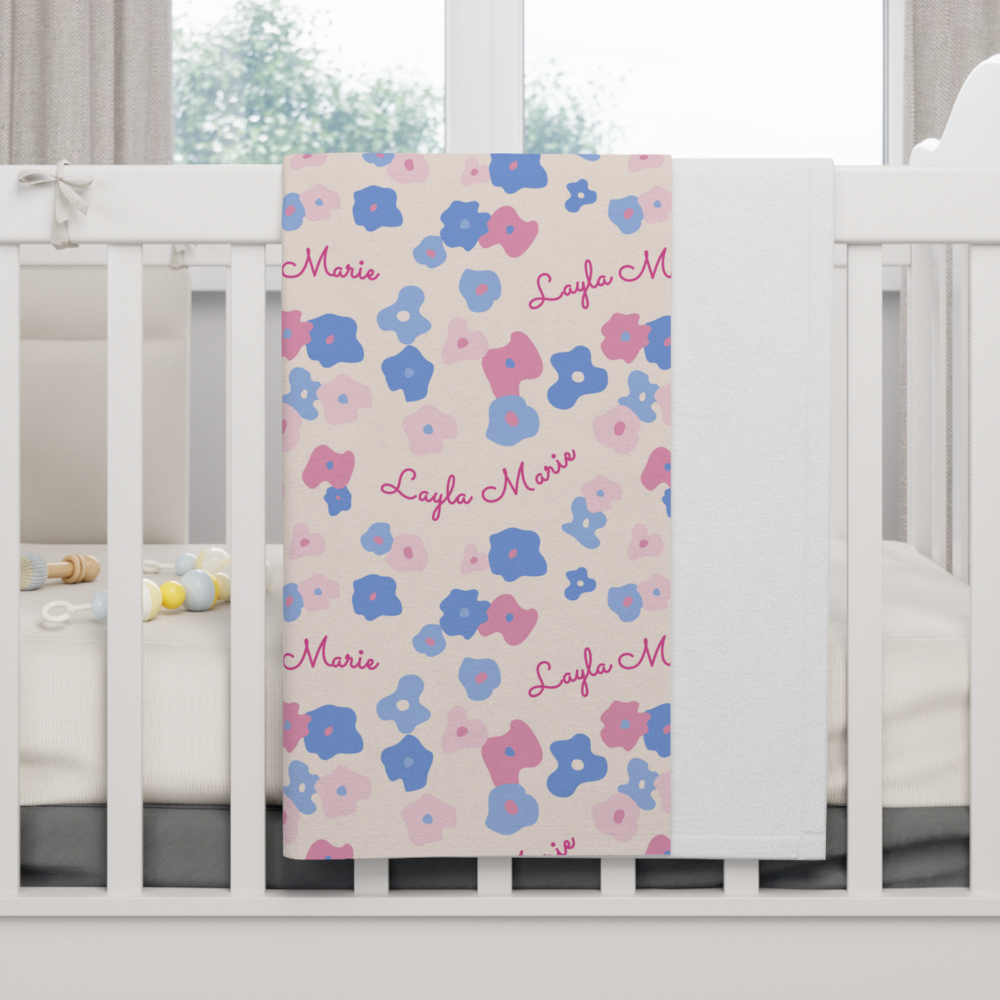 Fleece personalized baby blanket in graphic pink and blue daisy pattern hung over side of white crib with window in the background