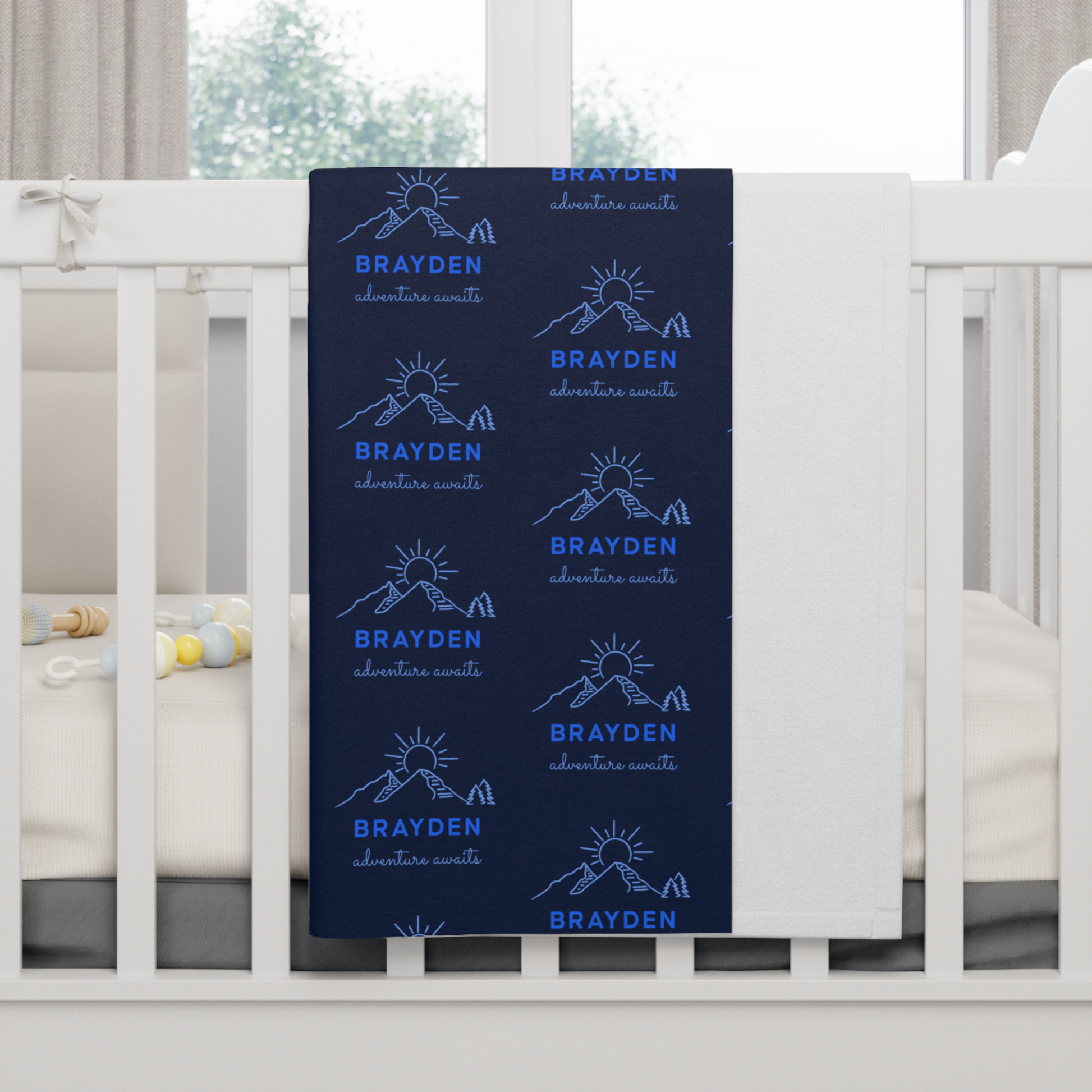Fleece personalized baby blanket in mountain adventure pattern hung over side of white crib with window in the background