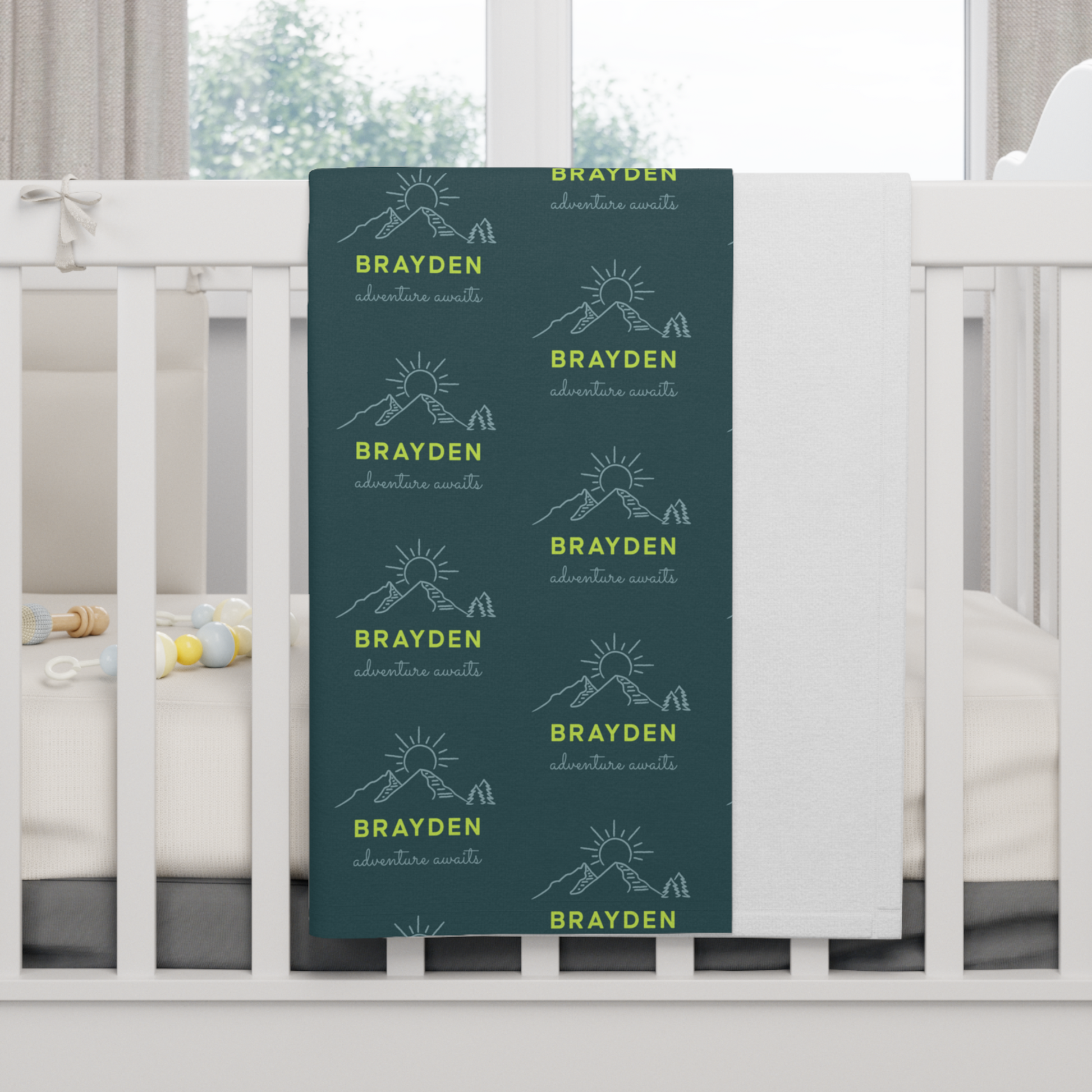 Fleece personalized baby blanket in green mountain adventure pattern hung over side of white crib with window in the background