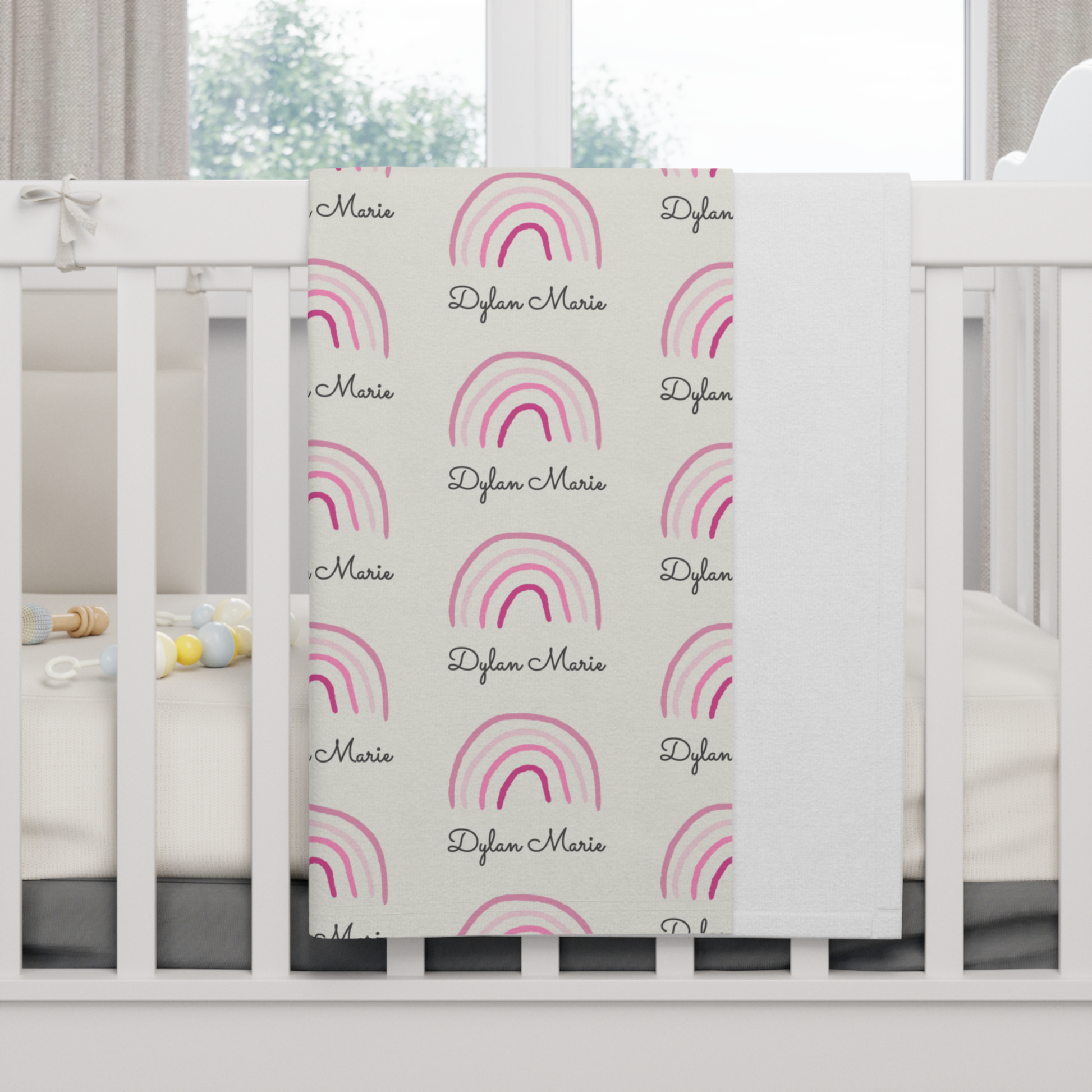 Fleece personalized baby blanket in pink rainbow pattern hung over side of white crib with window in the background