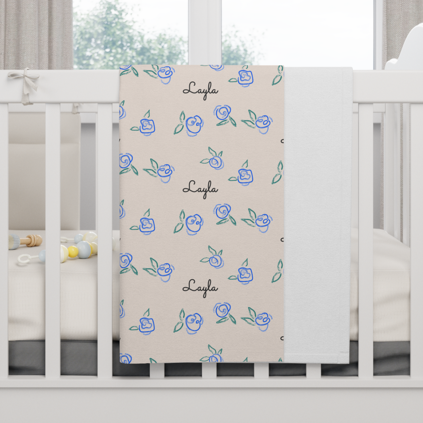 Fleece personalized baby blanket in blue rose pattern hung over side of white crib with window in the background