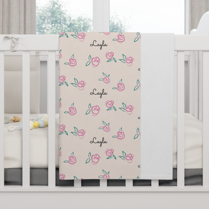 Fleece personalized baby blanket in pink rose with green leaves pattern hung over side of white crib with window in the background
