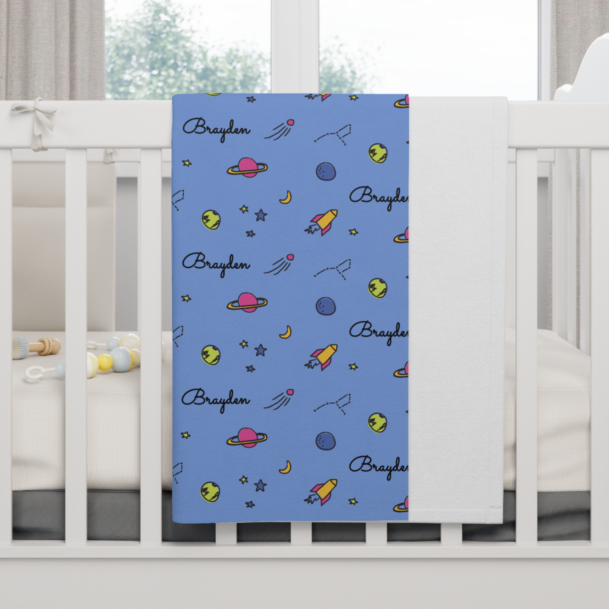 Fleece personalized baby blanket in space, rocket and stars pattern hung over side of white crib with window in the background